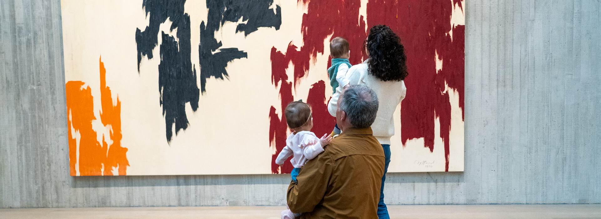 A grandfather sits holding his granddaughter while his daughter-in-law holds her son and they all look at a large abstract painting with black, red, orange, and bare canvas