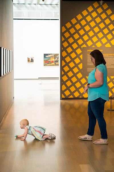 A baby girl crawls on the floor of an art gallery while her mom follows her