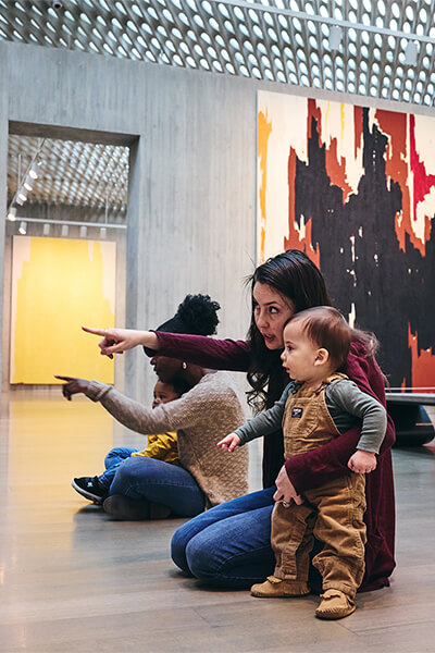 Two women and their babies look and point at art