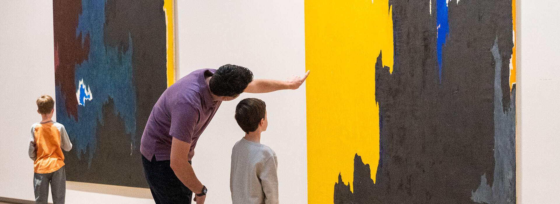 A father points at a painting with his two young sons next to him