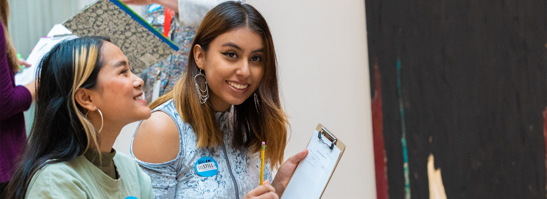One teen girl holds a clipboard and smiles while another smiles and looks up in an art gallery