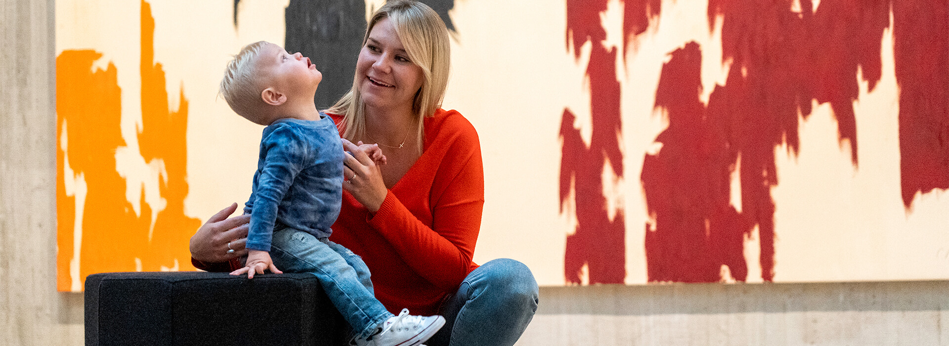 A mom sits with her toddler son in front of a big painting while he looks up at the ceiling in amazement