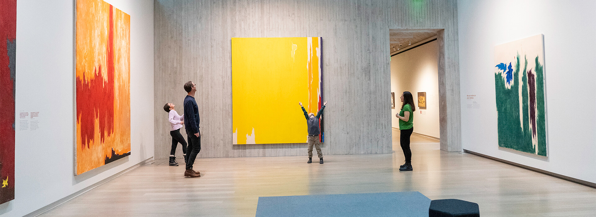 A family looks at colorful abstract art in a gallery while a young boy raises his arms up in the air