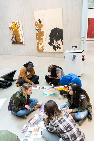 A group of students sit on the floor in a circle of an art gallery with a teacher