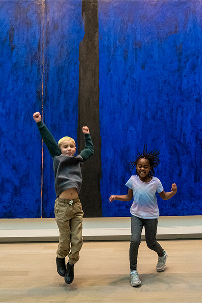 A boy and girl jump with excitement in front of a big blue abstract painting