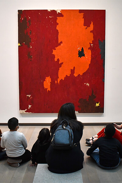 A group of students and teachers sit on the floor and a bench and look up at a big red abstract painting