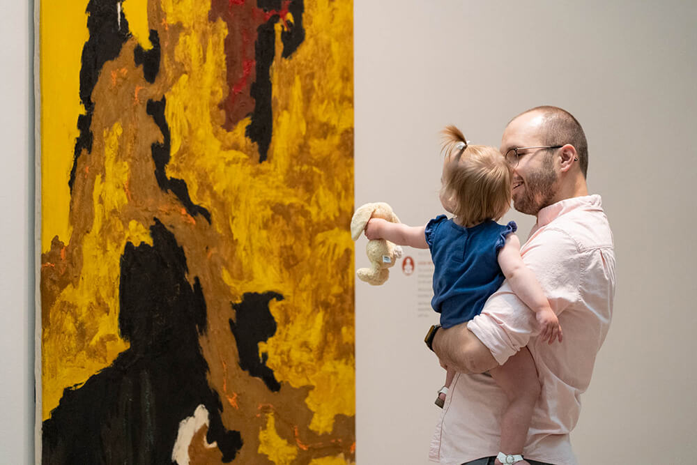 A dad holds his daughter up while she looks at a painting