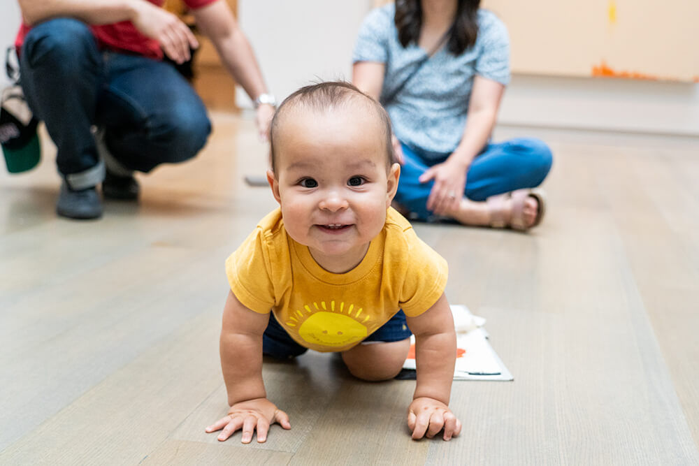 A baby smiles and looks at the camera while crawling on the floor