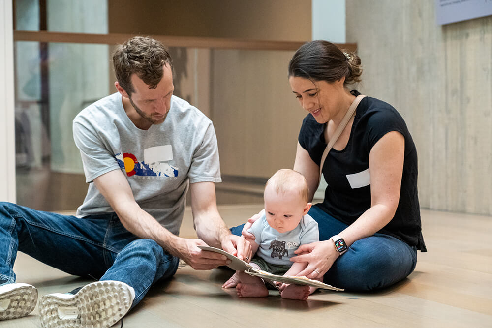 A mother and father sit on the floor with their baby as it looks at a book