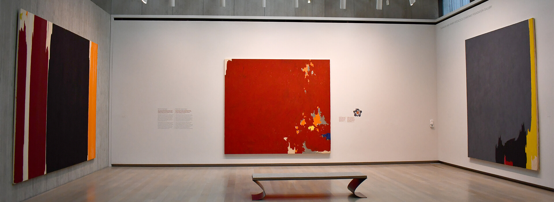 Gallery installation image with three large colorful abstract paintings and a bench