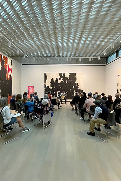 People sit in seats listening to a guitar player in an art gallery