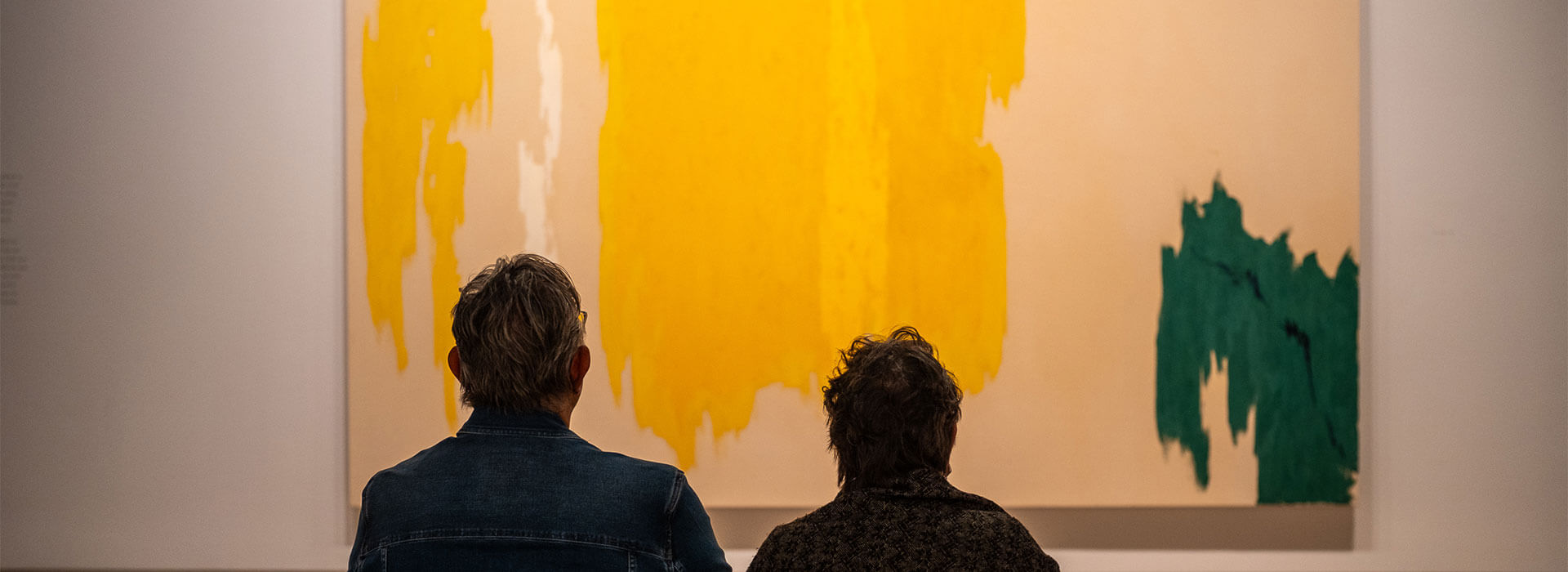 A man and woman sit next to each other on a bench and look at a large yellow, white, green, and tan abstract painting on the wall