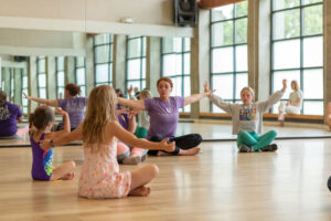 A teacher sits on the floor and instructs young students also sitting on the floor in a dance studio