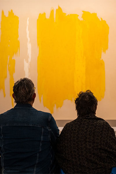 A man and woman sit next to each other on a bench and look at a large yellow, white, green, and tan abstract painting on the wall