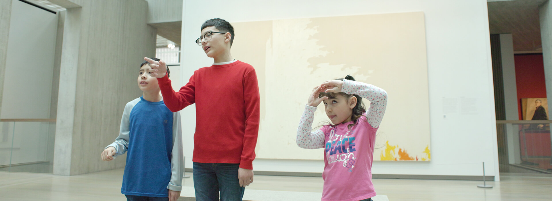 Three children look at a painting in a gallery, the boys point while the girl makes a circle shape with her hands on her forehead
