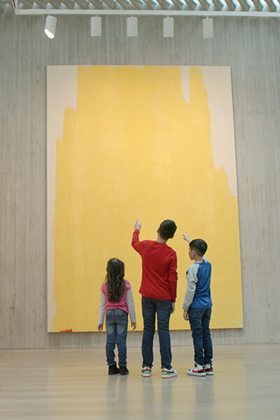 Three children look up at a tall abstract yellow painting on a concrete wall
