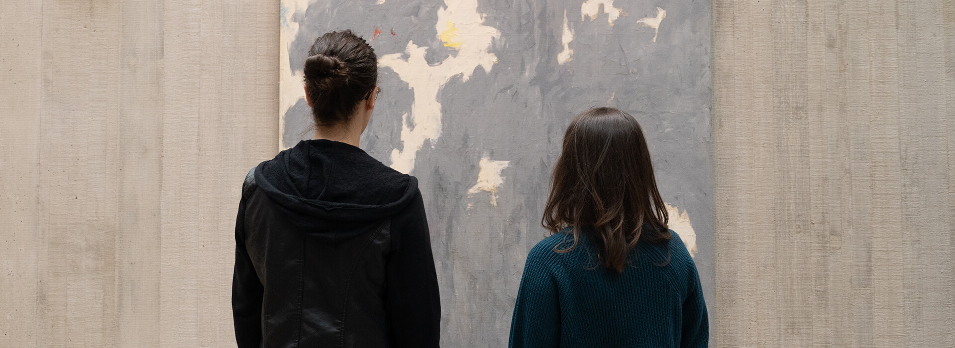 Two women with their backs to the camera look at a blue gray abstract painting on a concrete wall
