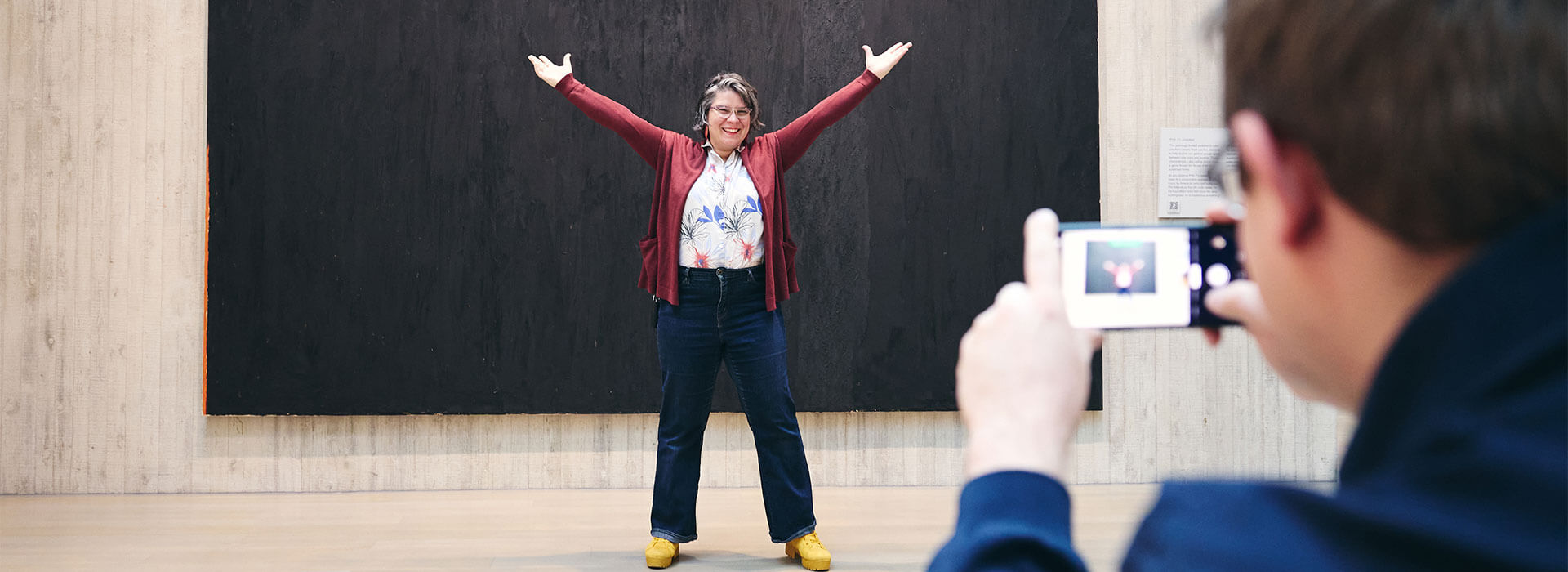 A man takes a photo of his wife in front of a big black abstract painting with his phone while she sticks her arms up in the air