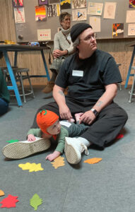 A man sits on the floor with his baby who is wearing a pumpkin hat