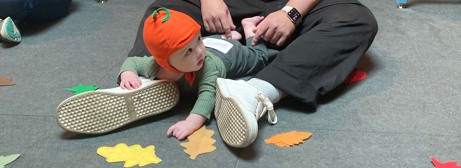 A man sits on the floor with his baby who is wearing a pumpkin hat