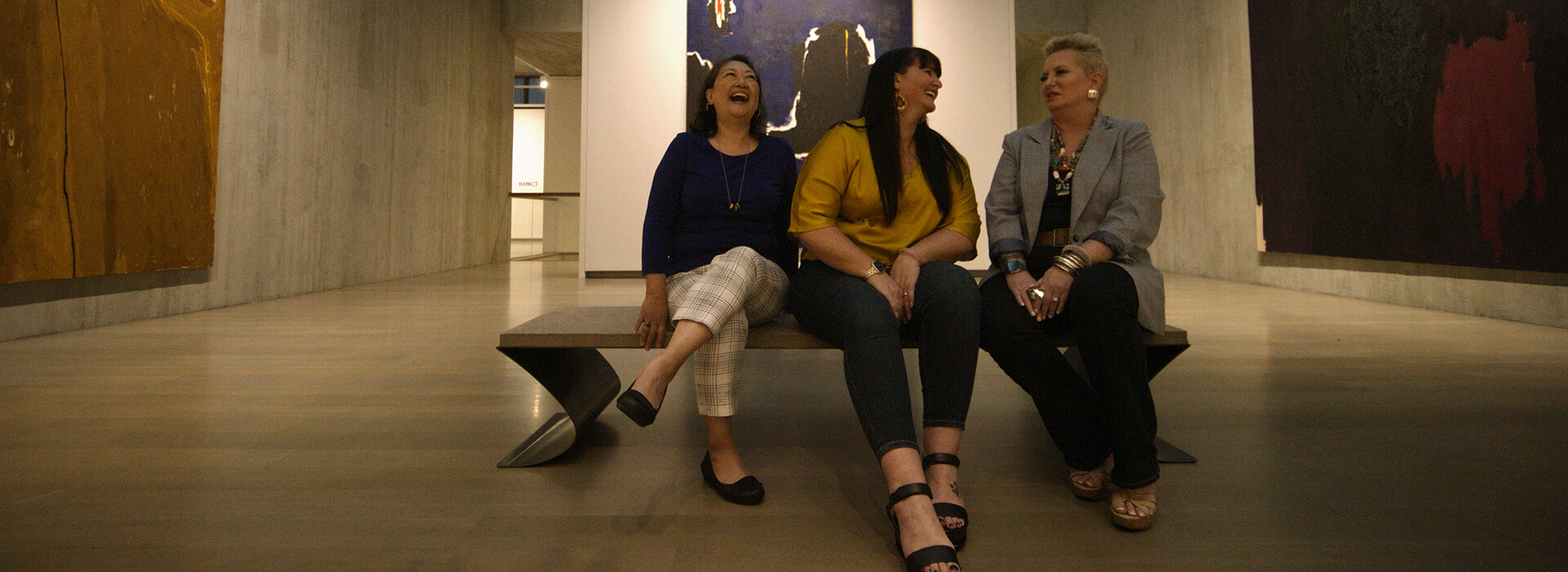 Three women sit in a dark gallery and laugh