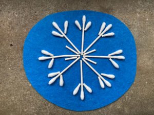 A blue piece of felt has a snowflake shaped art piece on it made out of cotton swabs