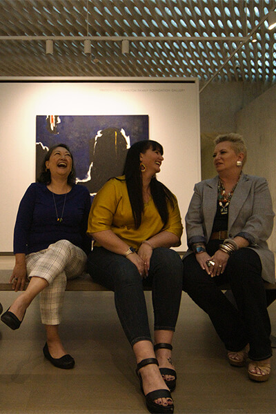 Three women sit in a dark gallery and laugh