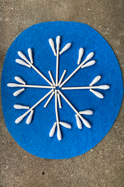 A blue piece of felt has a snowflake shaped art piece on it made out of cotton swabs