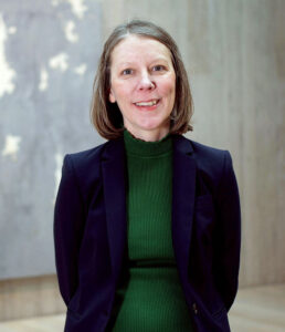 A woman wearing a green shirt and blue jacket smiles and stands in front of an abstract light blue gray painting on a concrete wall