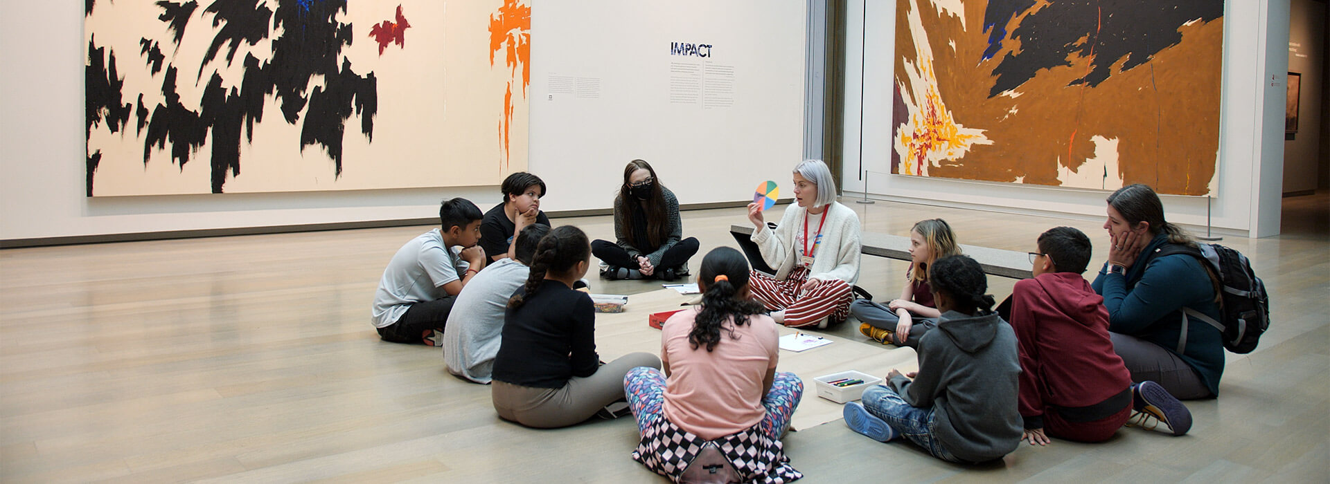 A group of students sit in a circle on the floor of a gallery while a teacher holds up a color wheel