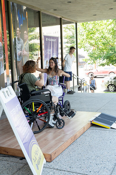 Two people in wheelchairs dance on a stage in front of a museum