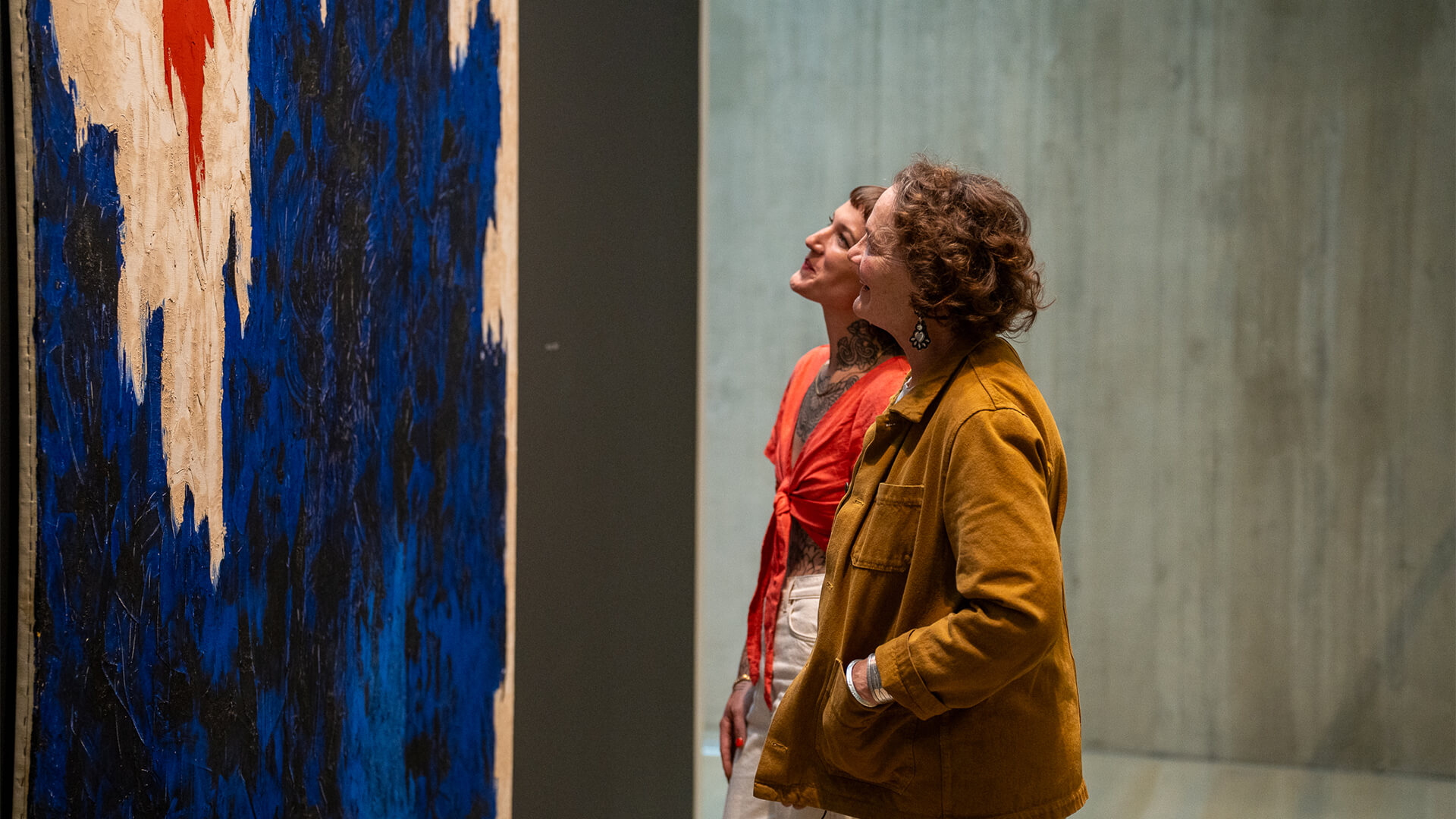 Two women look up at an abstract blue, white, black and orange painting and smile