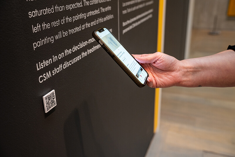 A woman's hand holds a phone and takes a picture of a QR code on a black wall