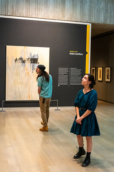 A woman looks up while a man behind her looks to the side with a partially treated painting behind them