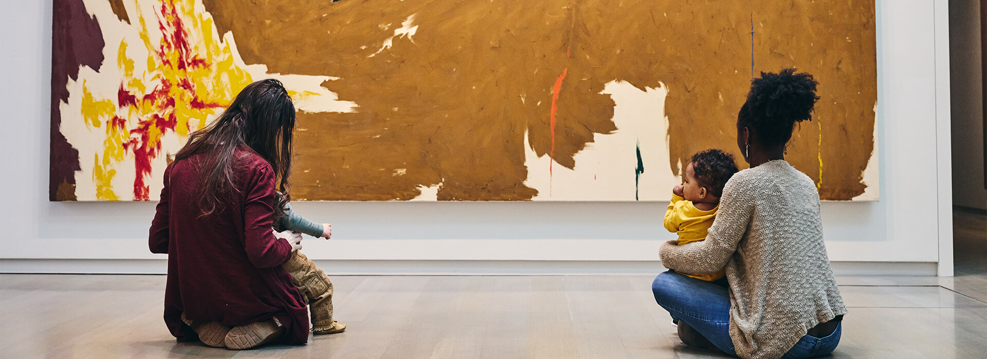 Two moms and their infants sit on the floor in front of a large abstract painting