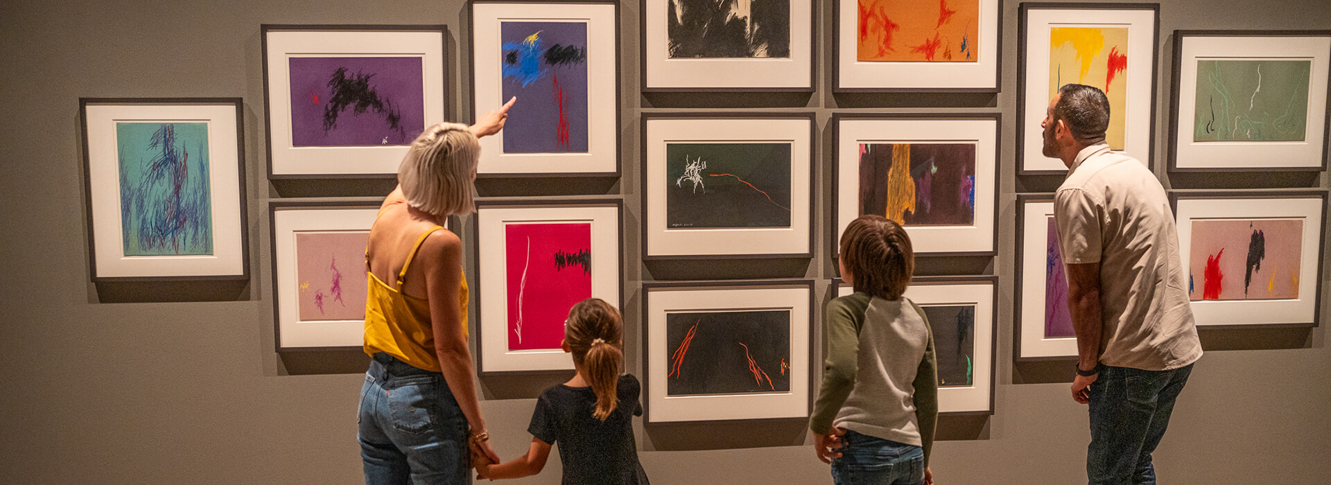 A family looks closely at a wall of framed pastel on construction paper artworks
