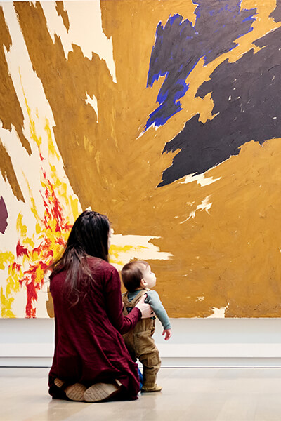 A mom and her infant son sit on the floor in front of a large abstract painting