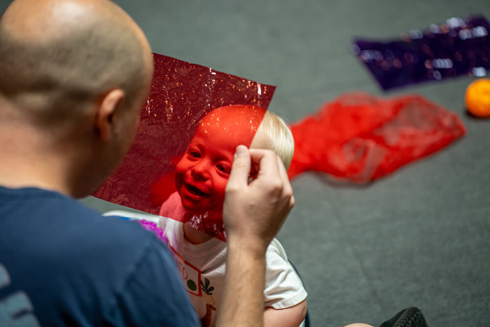 A father holds up a transparent piece of red plastic and looks through it as his baby looks through the other side, smiling back at him
