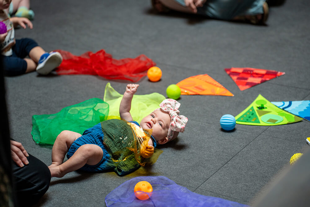 A baby girl wearing a bow on her head lays on the floor surrounded by colorful scarves and balls