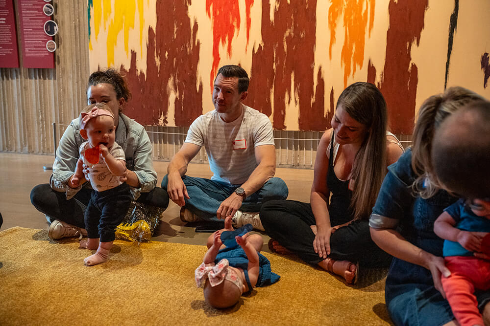 A group of caregivers sit with their babies on a yellow carpet in an art gallery