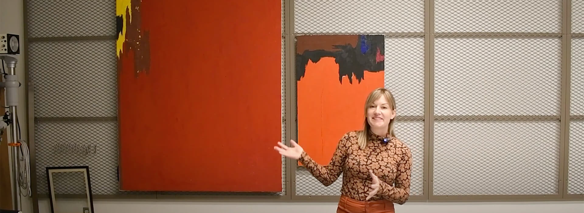 Bailey Placzek stands in front of two abstract paintings hanging on wire panels and gestures toward one.
