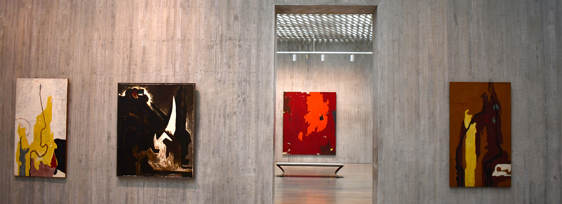 Three abstract paintings hang on a concrete wall and one hangs on a wall visible through a doorway in between the paintings