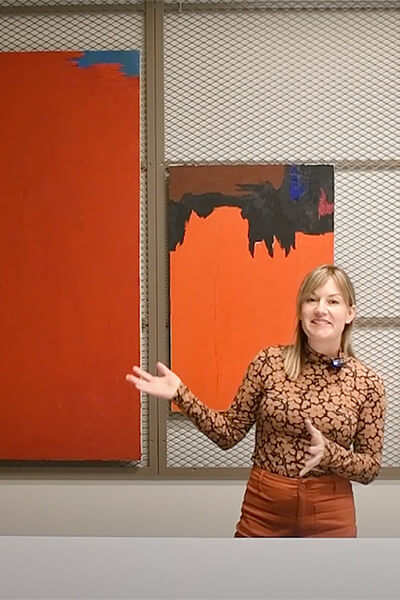 Bailey Placzek stands in front of two abstract paintings hanging on wire panels and gestures toward one.