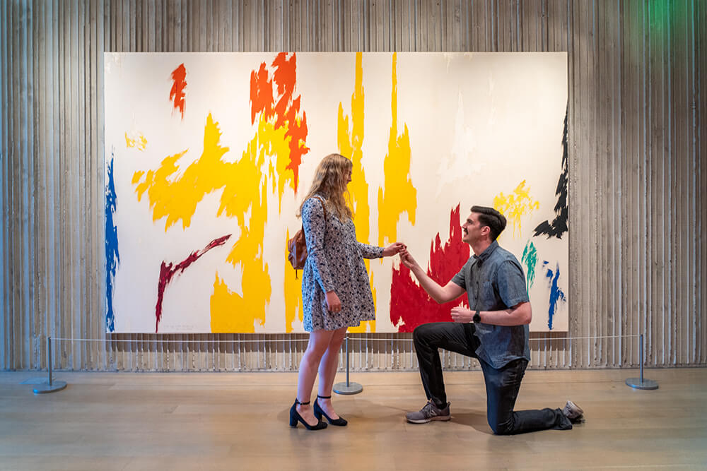 A man proposes to a woman in front of a large colorful painting