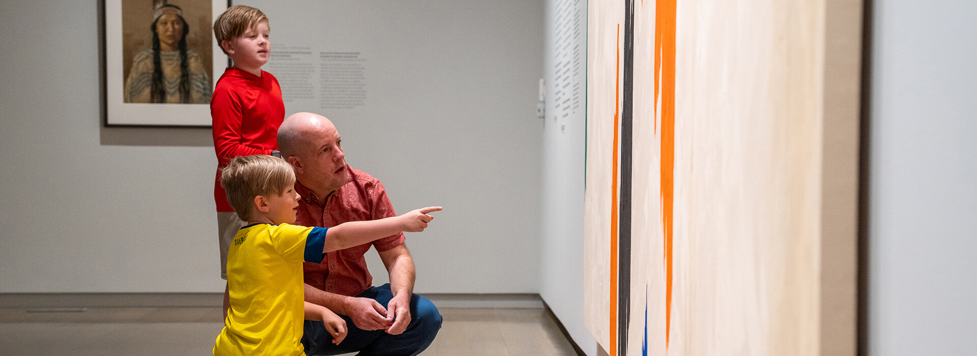 A dad kneels down next to one of his sons while the son points at a painting, the other son stands behind them