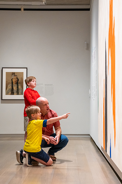 A dad kneels down next to one of his sons while the son points at a painting, the other son stands behind them