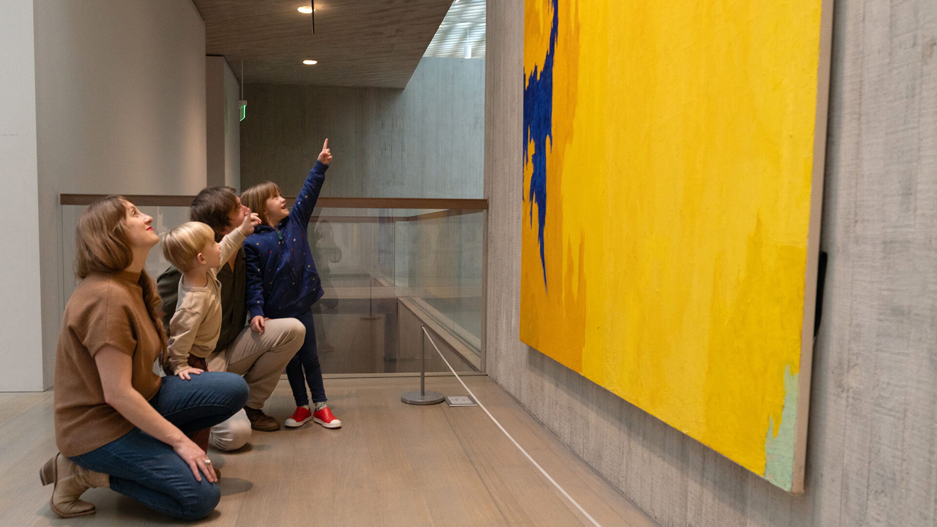 A family kneels on the ground and looks up at a large yellow abstract painting