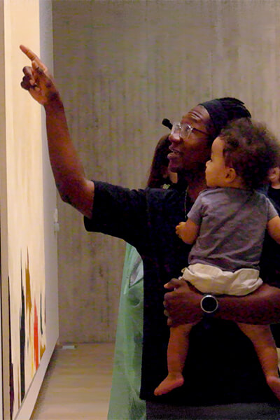 A father holds his infant son and points up at an artwork