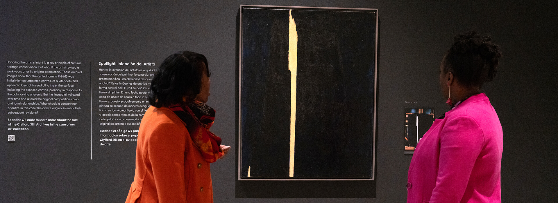 Two women wearing brightly colored jackets look at a dark painting with a vertical yellow line on a black wall