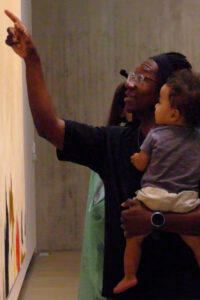 A father holds his baby and points up at some art while the baby looks at it.